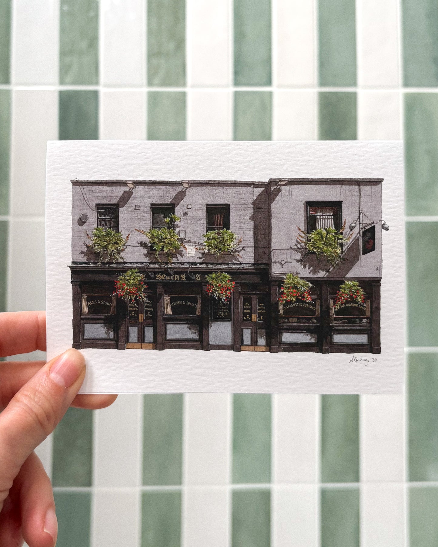 A hand holds the horizontal A6 greeting card of THE SEVEN STARS in Holborn against a background of sage green and white tiles, highlighting the vibrant watercolor flower baskets.