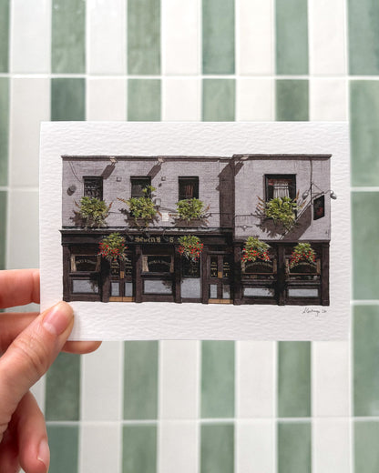 A hand holds the horizontal A6 greeting card of THE SEVEN STARS in Holborn against a background of sage green and white tiles, highlighting the vibrant watercolor flower baskets.