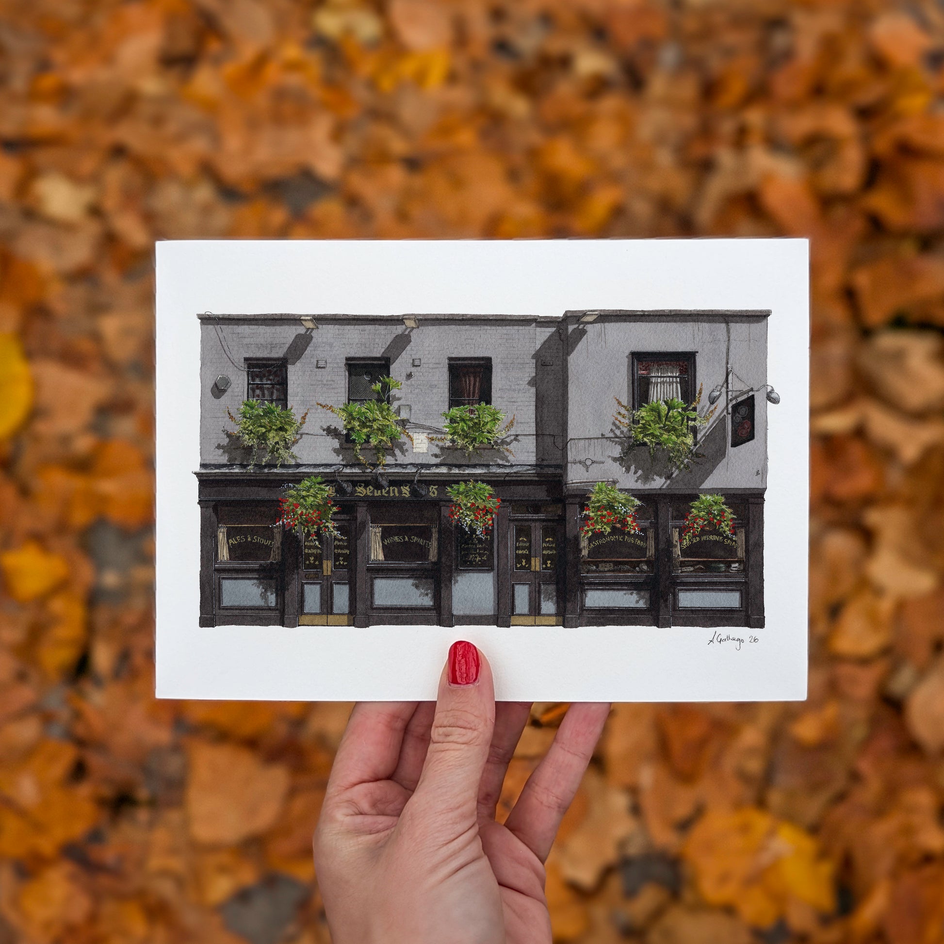 A hand with red nail polish holds the horizontal art print of THE SEVEN STARS in Holborn by Ana Gallego against a seasonal background of fallen orange and brown autumn leaves.