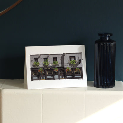 A horizontal greeting card of THE SEVEN STARS in Holborn stands on a white mantelpiece against a deep navy blue wall, next to a textured blue glass vase.