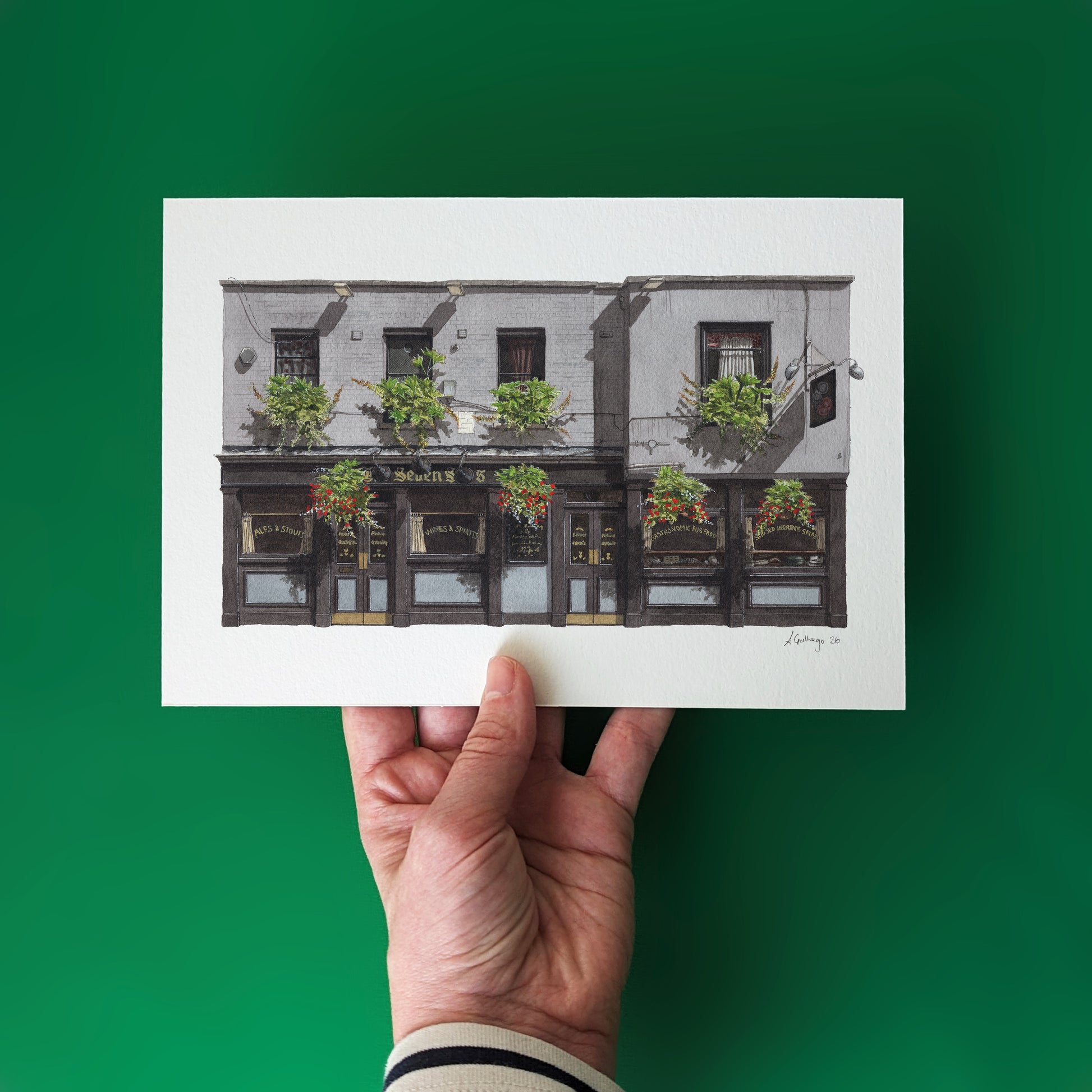 A hand holds the white art print of THE SEVEN STARS in Holborn against a solid, deep emerald green background, highlighting the vibrant green flower baskets in the watercolor illustration.