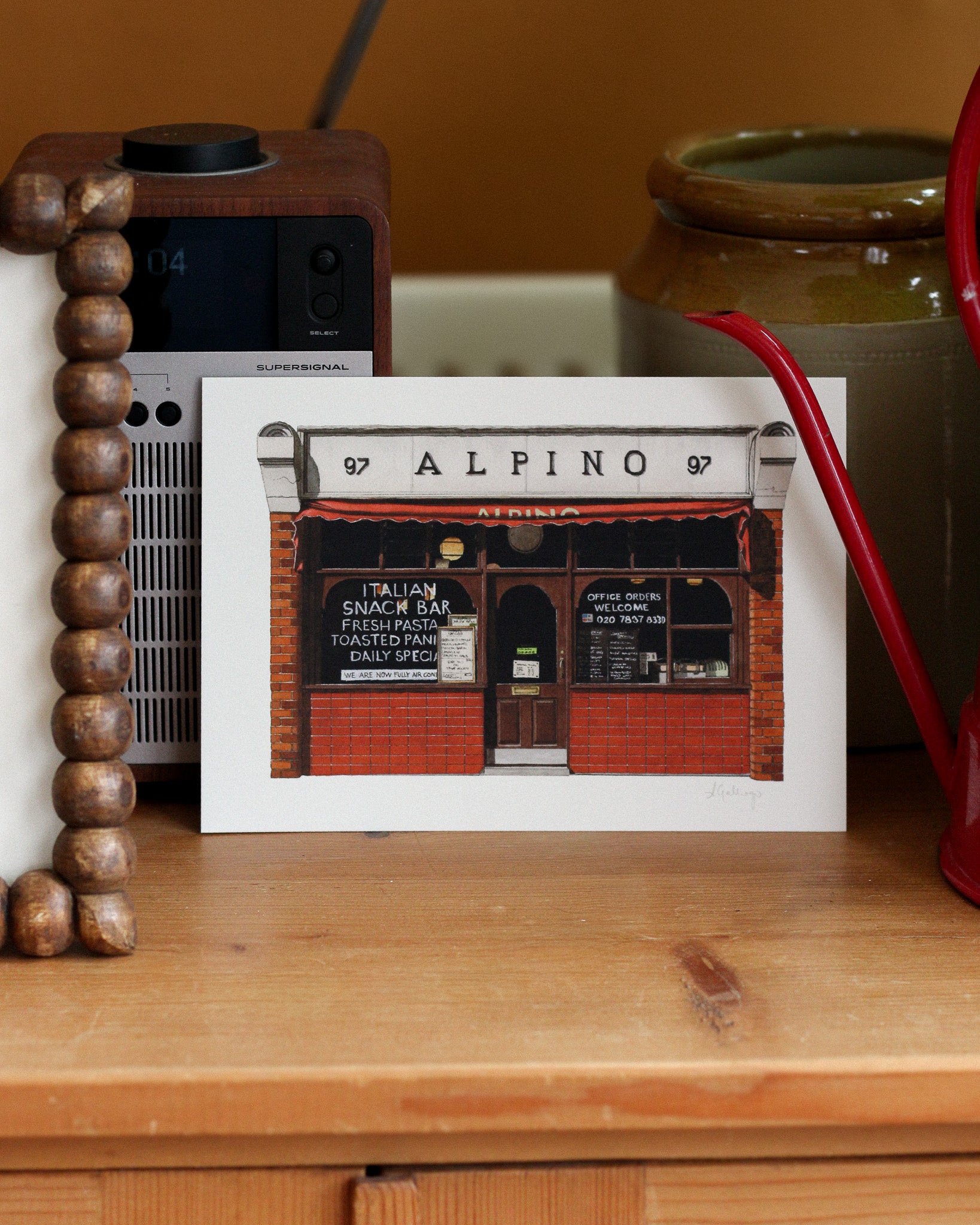A professional art print of Alpino, an iconic Italian snack bar with red brick and a striped awning, displayed on a wooden shelf next to a vintage radio and ceramic jars.