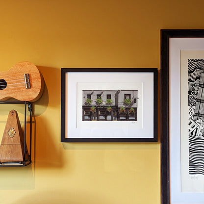 A watercolor print of THE SEVEN STARS in Holborn is displayed in a black frame with a white mount, hanging on a warm mustard-yellow wall next to a wooden ukulele and a metronome.