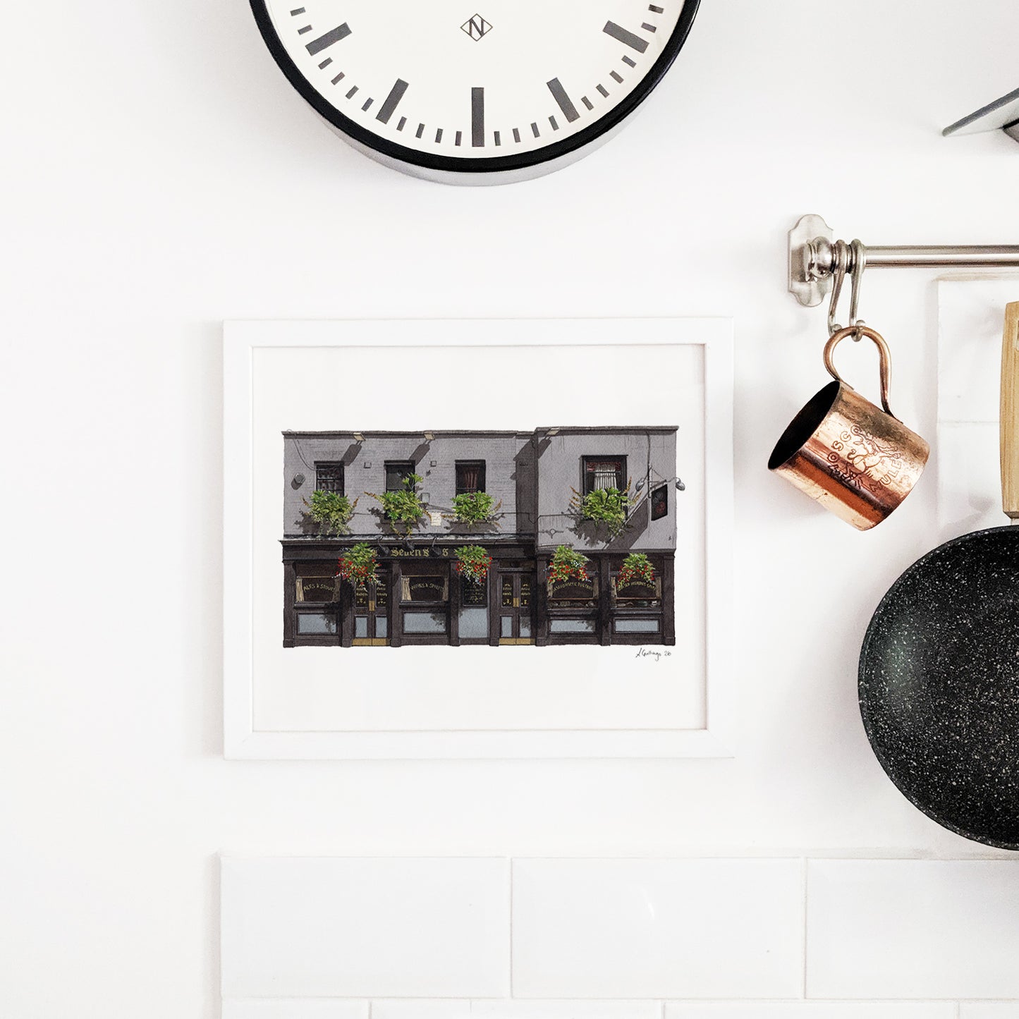 A white-framed art print of THE SEVEN STARS in Holborn hangs on a white tiled kitchen wall, styled alongside a copper mug, a black frying pan, and a minimalist wall clock.