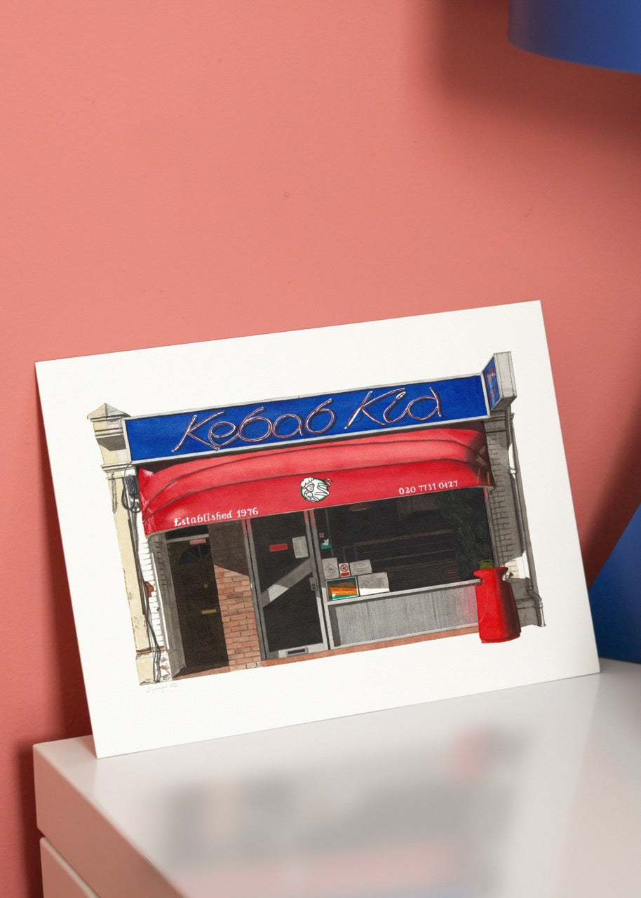 Framed artwork of a store front with a red awning and blue sign on a white surface.