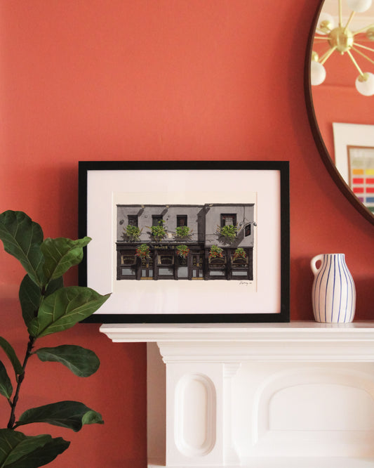 The illustration of THE SEVEN STARS in Holborn is shown in a black frame with a white mat, resting on a white fireplace mantel against a bold terracotta pink wall next to a Fiddle Leaf Fig plant.