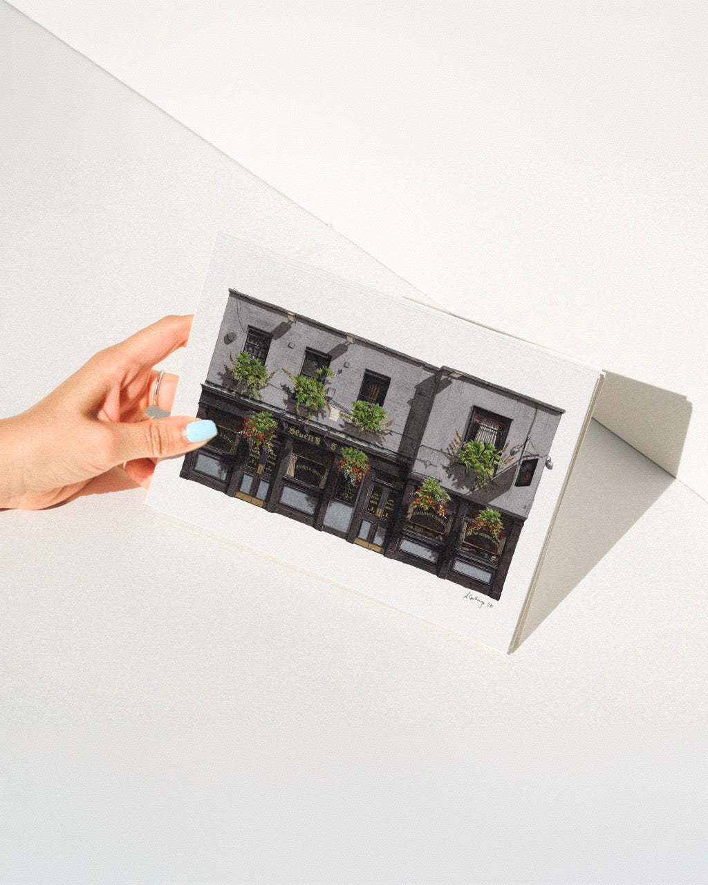 A hand with light blue nail polish holds the horizontal watercolor print of THE SEVEN STARS in Holborn in bright, direct sunlight, casting a sharp shadow against a minimalist white studio background.