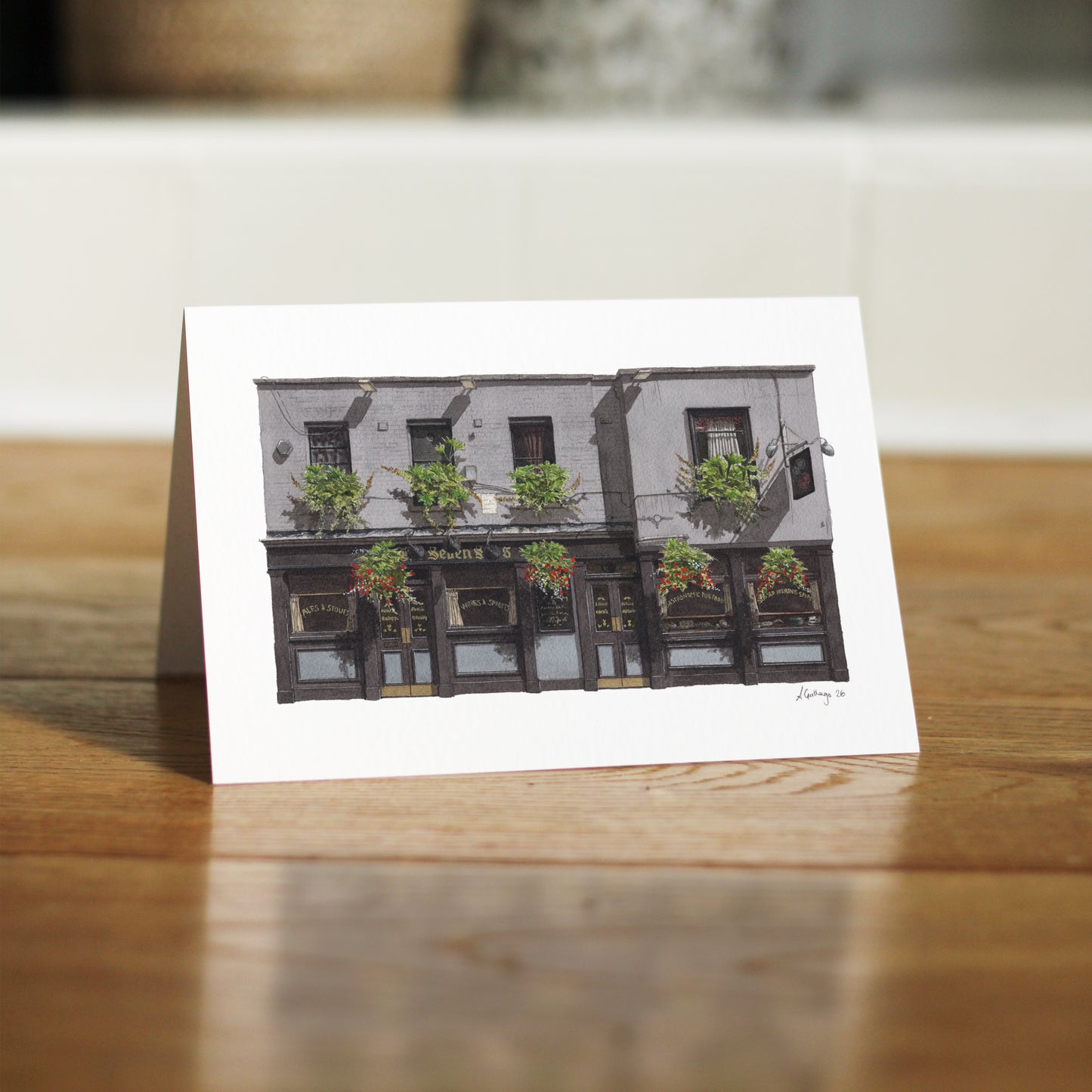 A close-up, low-angle shot of THE SEVEN STARS in Holborn greeting card standing independently on a polished wooden floor, showing the card's sturdy fold and matte finish.
